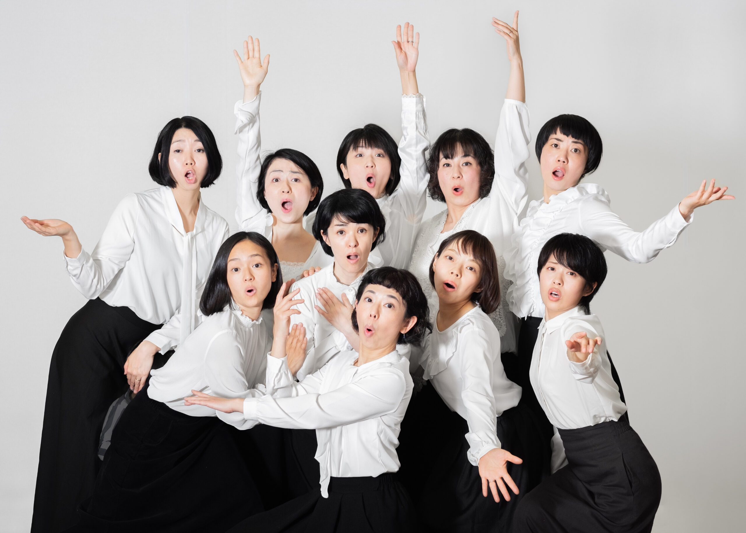 白いブラウス、黒いスカートを着た10名の女性が集まってこちらを見ている。思い思いに手を伸ばすなどポーズをとっており、驚いたような口のあいた表情をしている。Ten women dressed in white blouses and black skirts face the camera, striking expressive poses. With mouths open in surprise and arms reaching out in various directions, they convey a sense of dramatic tension.