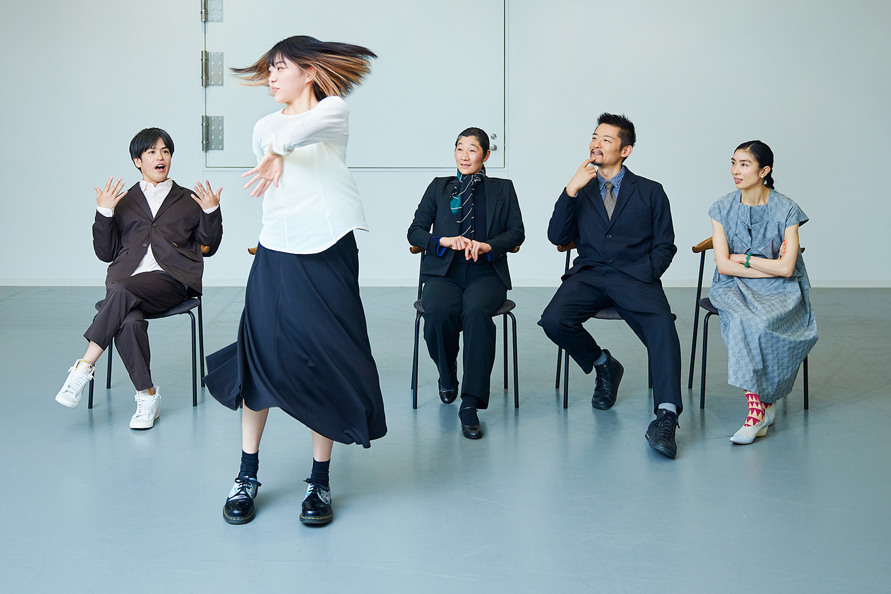 白いシャツと黒いスカートを着た女性出演者が勢いよく回転して踊っている様子。その背後には椅子に座った4人の出演者が審査員風の衣装を着て横一列に座っており、それぞれ異なる表情で彼女の動きを見つめている。スタジオのようなシンプルな白い室内。